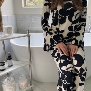 May include: A long-sleeved, two-piece outfit featuring a cream background with large black floral patterns. The outfit has a round neckline and flared sleeves. The matching pants have a similar floral design. The outfit is suitable for lounging or casual wear.