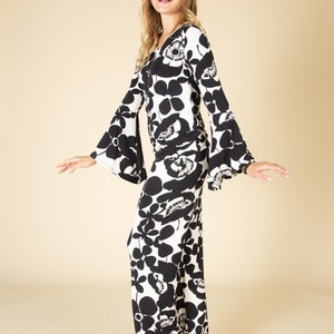 May include: A long, flowing jumpsuit with a black and white floral pattern. The jumpsuit features a V-neckline and flared sleeves. The model is smiling and wearing the jumpsuit in a studio setting.