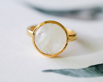 May include: A gold-colored ring featuring a large, round, white gemstone. The gemstone has a milky, opaque appearance and is set in a gold bezel. The ring band is also gold. The ring is displayed on a white surface with a subtle floral pattern.