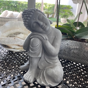 May include: A gray Buddha statue in a seated pose, with eyes closed and head resting on a knee. The statue has a textured surface and is placed on a dark, woven surface. The statue is a decorative item.