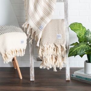 May include: Three decorative throws with fringe are displayed. One is striped, one has a chevron pattern, and the third has a striped pattern. All are cream-colored with fringe. The throws are draped over a white ladder and a small stool. Each throw has a "NINE ZEN" label.