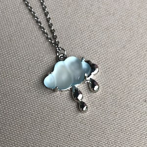 May include: A silver-toned necklace featuring a cloud pendant with a light blue, pearlescent center and silver raindrops. The cloud pendant is approximately 1 inch wide and hangs from a delicate chain.