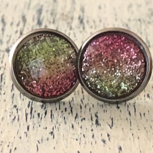 May include: A pair of round stud earrings featuring a metallic silver-toned setting and a domed surface with a gradient glitter finish that transitions from a vibrant magenta pink to a bright lime green across the circular face of each earring.