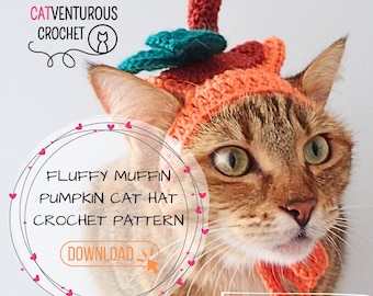May include: A brown tabby cat wearing a crocheted pumpkin hat with a green stem. The hat is orange and brown with a fluffy texture. The text "FLUFFY MUFFIN PUMPKIN CAT HAT CROCHET PATTERN DOWNLOAD" is in a circle with pink hearts around it. The text "#fashionablycatventurous" is at the bottom of the image.