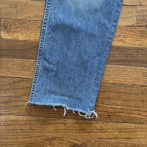 May include: Close-up of the bottom of a pair of blue jeans with a frayed hem. The denim is a medium wash, and the frayed edge has white threads. The jeans are on a wooden surface.