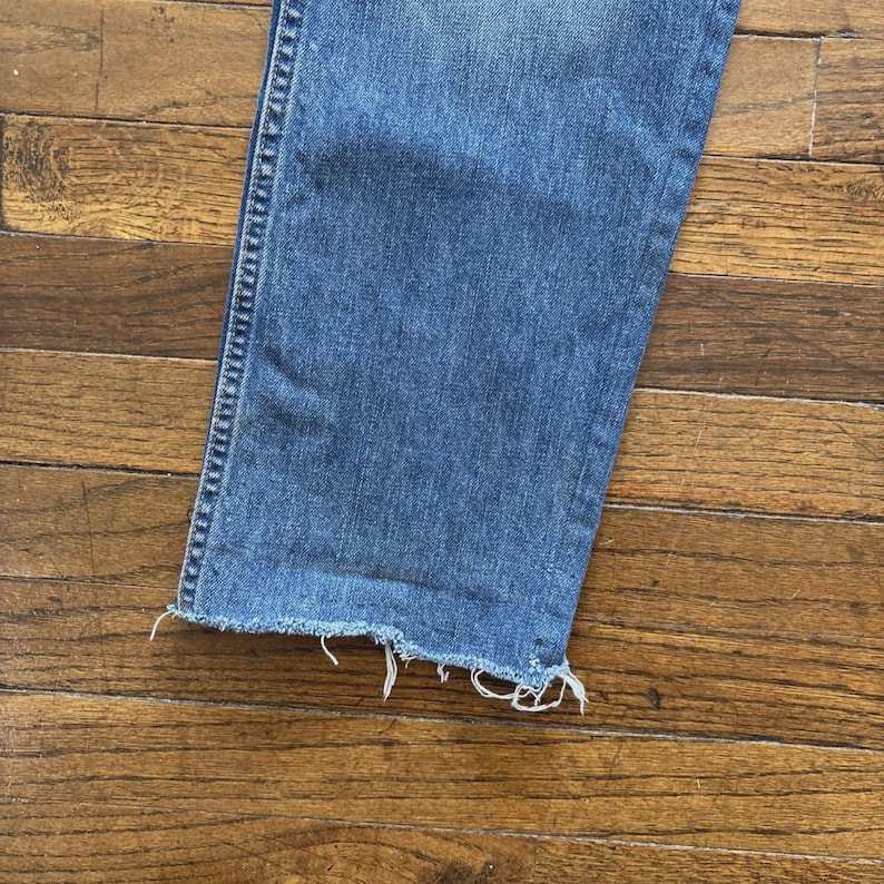 May include: Close-up of the bottom of a pair of blue jeans with a frayed hem. The denim is a medium wash, and the frayed edge has white threads. The jeans are on a wooden surface.