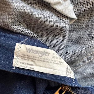 Puede incluir: Primer plano de unos vaqueros de mezclilla azules con una etiqueta Wrangler. La etiqueta incluye instrucciones de lavado y dice "Made in USA" y "100% COTTON". Los vaqueros tienen una cremallera. La etiqueta tambi&eacute;n indica la talla de los vaqueros.