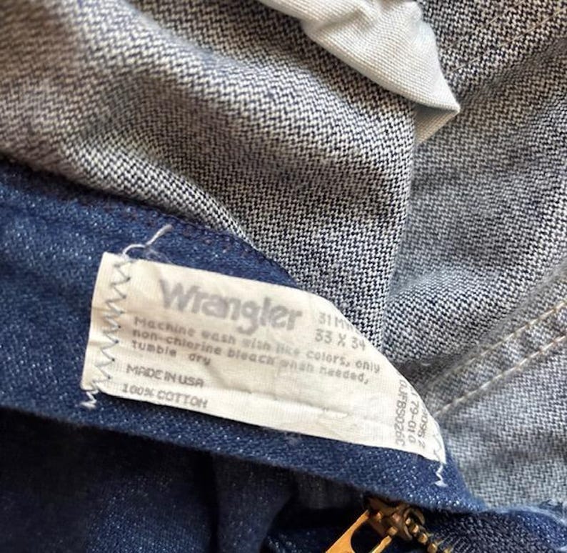 Puede incluir: Primer plano de unos vaqueros de mezclilla azules con una etiqueta Wrangler. La etiqueta incluye instrucciones de lavado y dice "Made in USA" y "100% COTTON". Los vaqueros tienen una cremallera. La etiqueta tambi&eacute;n indica la talla de los vaqueros.
