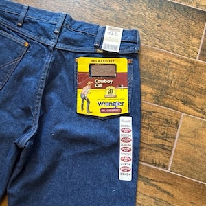 Puede incluir: Primer plano de unos vaqueros azules Wrangler Cowboy Cut de corte relajado. Los vaqueros tienen una etiqueta amarilla y roja en el bolsillo trasero con el logotipo de Wrangler y la talla 31. Varias etiquetas de talla, 33x34, est&aacute;n adheridas a los vaqueros.