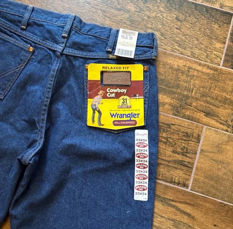 Puede incluir: Primer plano de unos vaqueros azules Wrangler Cowboy Cut de corte relajado. Los vaqueros tienen una etiqueta amarilla y roja en el bolsillo trasero con el logotipo de Wrangler y la talla 31. Varias etiquetas de talla, 33x34, est&aacute;n adheridas a los vaqueros.