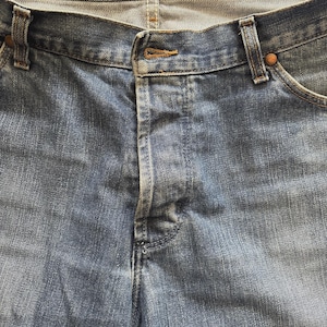 May include: Close-up of blue denim jeans. The jeans have a button closure and a zipper fly. The fabric shows a faded wash, with visible stitching and rivets. The jeans are a classic style.