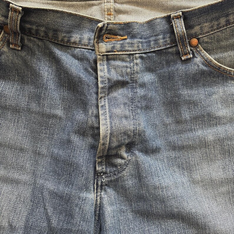 May include: Close-up of blue denim jeans. The jeans have a button closure and a zipper fly. The fabric shows a faded wash, with visible stitching and rivets. The jeans are a classic style.