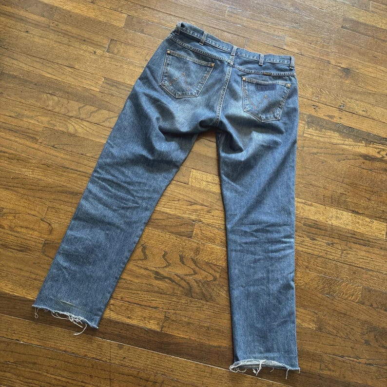 May include: Light blue denim jeans with a classic five-pocket design. The jeans have a straight leg cut and a distressed hem. The back pockets feature a decorative stitch pattern. The jeans are laid out on a wooden surface.