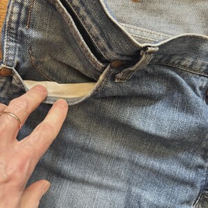 May include: Close-up of blue denim jeans with a visible pocket. The pocket lining is a light beige color. The jeans have copper-colored rivets and a button closure. The fabric shows signs of wear.