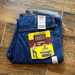 Puede incluir: Vaqueros Wrangler azul oscuro, corte Cowboy, talla 33x34, con etiqueta amarilla y roja. La etiqueta presenta una ilustraci&oacute;n de vaquero y el logotipo de Wrangler. Los vaqueros est&aacute;n doblados y expuestos sobre una superficie de madera.