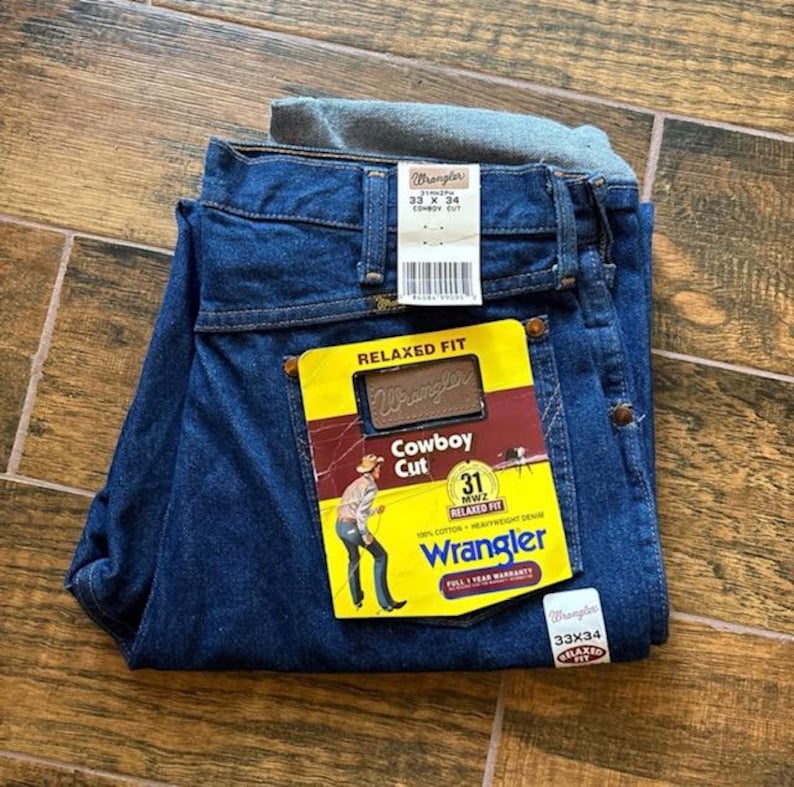 Puede incluir: Vaqueros Wrangler azul oscuro, corte Cowboy, talla 33x34, con etiqueta amarilla y roja. La etiqueta presenta una ilustraci&oacute;n de vaquero y el logotipo de Wrangler. Los vaqueros est&aacute;n doblados y expuestos sobre una superficie de madera.