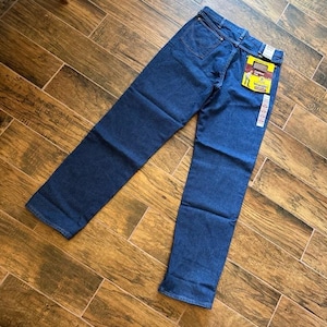 Puede incluir: Vaqueros de mezclilla azul oscuro con un corte recto cl&aacute;sico. Los vaqueros tienen un dise&ntilde;o tradicional de cinco bolsillos y una etiqueta de marca en el bolsillo trasero. Los vaqueros est&aacute;n colocados sobre una superficie de madera.