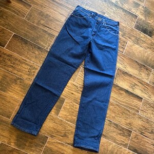 Puede incluir: Un par de vaqueros de mezclilla azul. Los vaqueros tienen bragueta con botones, dise&ntilde;o de cinco bolsillos y corte recto. La mezclilla es de lavado medio y los vaqueros est&aacute;n sobre una superficie de madera.