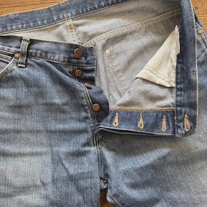 May include: Light blue denim jeans with a button fly and copper-colored buttons. The jeans are unbuttoned, revealing the interior construction and stitching details. The fabric shows signs of wear.