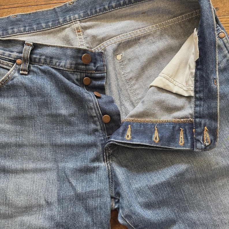 May include: Light blue denim jeans with a button fly and copper-colored buttons. The jeans are unbuttoned, revealing the interior construction and stitching details. The fabric shows signs of wear.