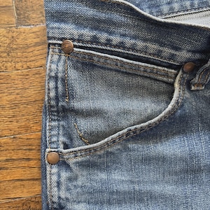 May include: Close-up of blue denim jeans, showing the pocket, rivets, and stitching details. The jeans have a classic design with a faded wash, and the rivets are a copper color.