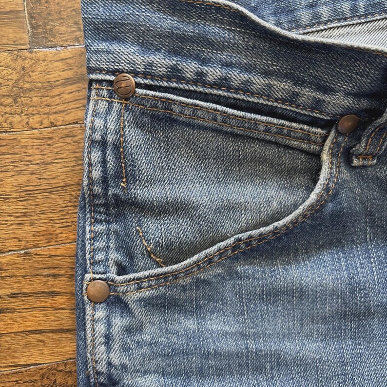 May include: Close-up of blue denim jeans, showing the pocket, rivets, and stitching details. The jeans have a classic design with a faded wash, and the rivets are a copper color.