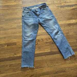 May include: Light blue denim jeans with a faded wash and distressed edges. The jeans have a classic five-pocket design and a straight leg fit. The jeans are laid flat on a wooden surface.