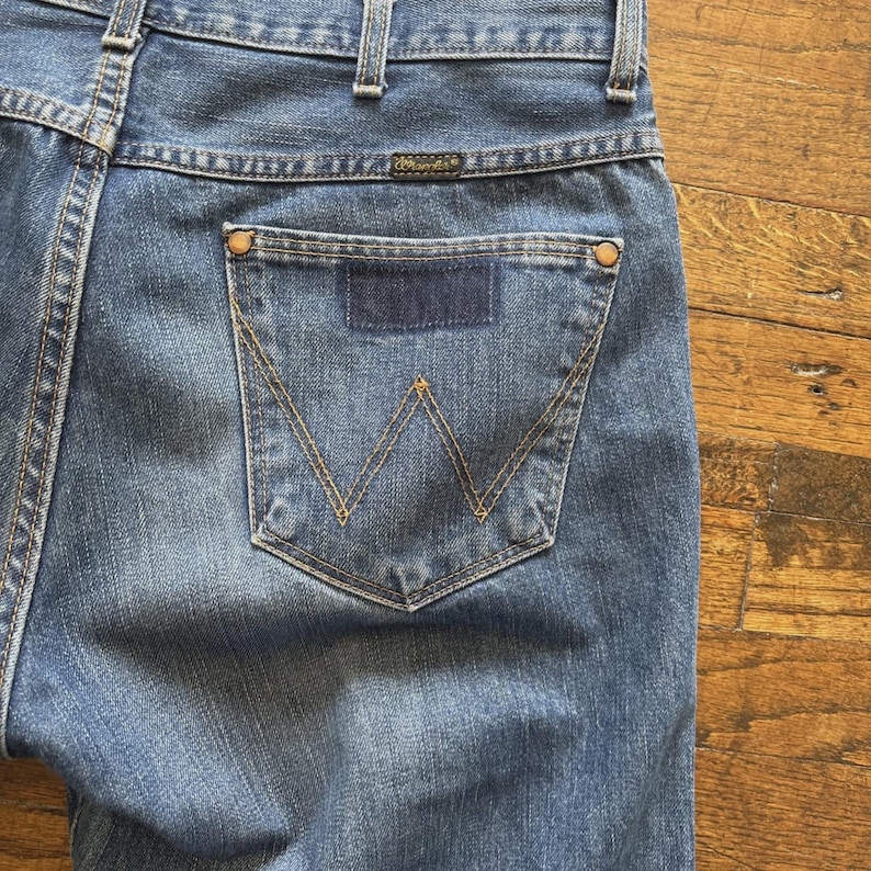 May include: Close-up of blue denim jeans with a Wrangler logo on the back pocket. The jeans have a classic five-pocket design with a dark blue patch and a signature "W" stitching on the back pocket.