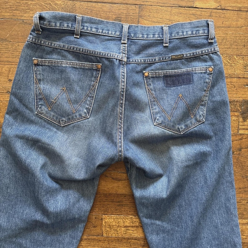 May include: Light blue denim jeans with the Wrangler logo on the back pocket. The jeans have a classic five-pocket design with a straight leg fit. The stitching is a contrasting tan color.