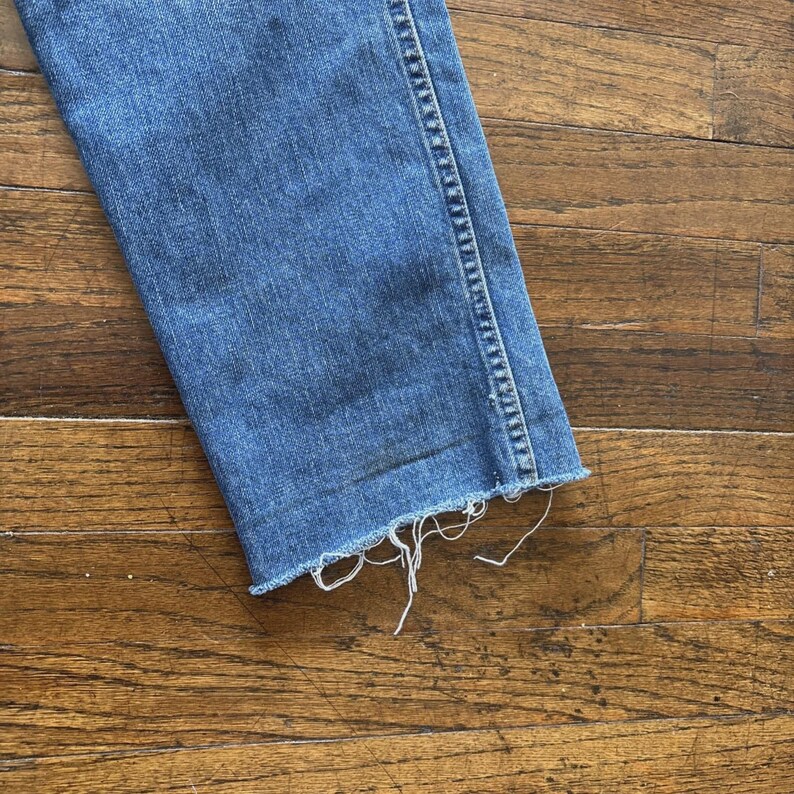 May include: Close-up of a section of blue denim jeans with a frayed, unfinished hem. The jeans are a medium wash and show signs of wear. The fabric is slightly faded, with visible stitching along the side seam.