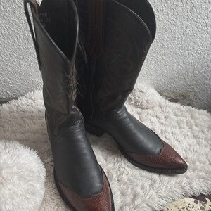 Könnte beinhalten: Cowboy-Stiefel mit schwarzen Lederoberteilen und braunen, strukturierten Zehenkappen. Die Stiefel haben dekorative Nähte und ein klassisches Western-Design.
