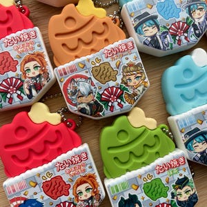 May include: A collection of colorful, taiyaki-shaped keychains. Each keychain features a different color, including red, orange, green, and blue, with a detailed illustration of anime characters and Japanese text. The keychains are attached to silver ball chains.