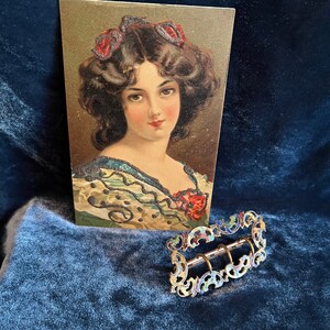 Op de afbeelding: Een vintage ansichtkaart met een vrouw met donker haar en een rode strik, naast een gouden gesp met blauwe, groene en rode emaille accenten.