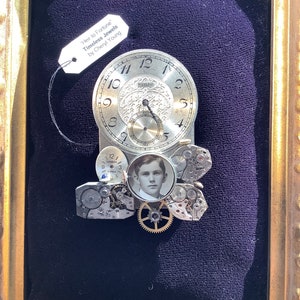 May include: A framed artwork featuring a clock face with gears and a small photo of a young man. The artwork is titled "Heir to Fortune Timeless Jewels by Cheryl Young".