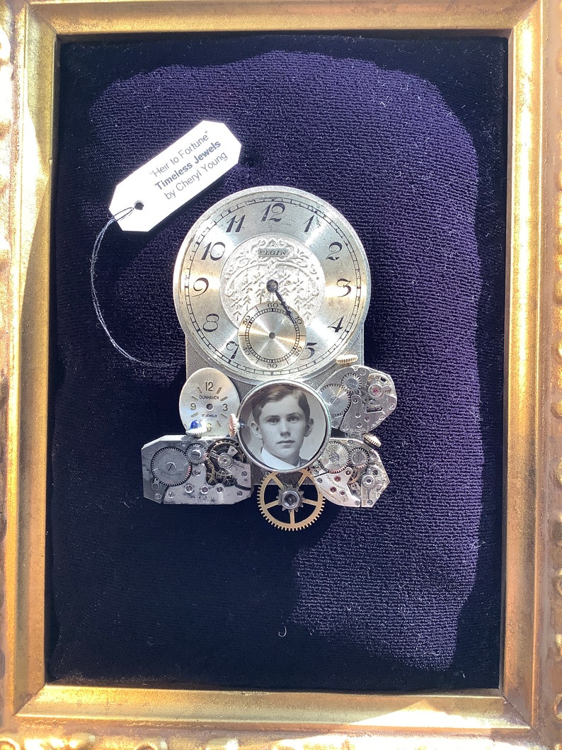 May include: A framed artwork featuring a clock face with gears and a small photo of a young man. The artwork is titled "Heir to Fortune Timeless Jewels by Cheryl Young".