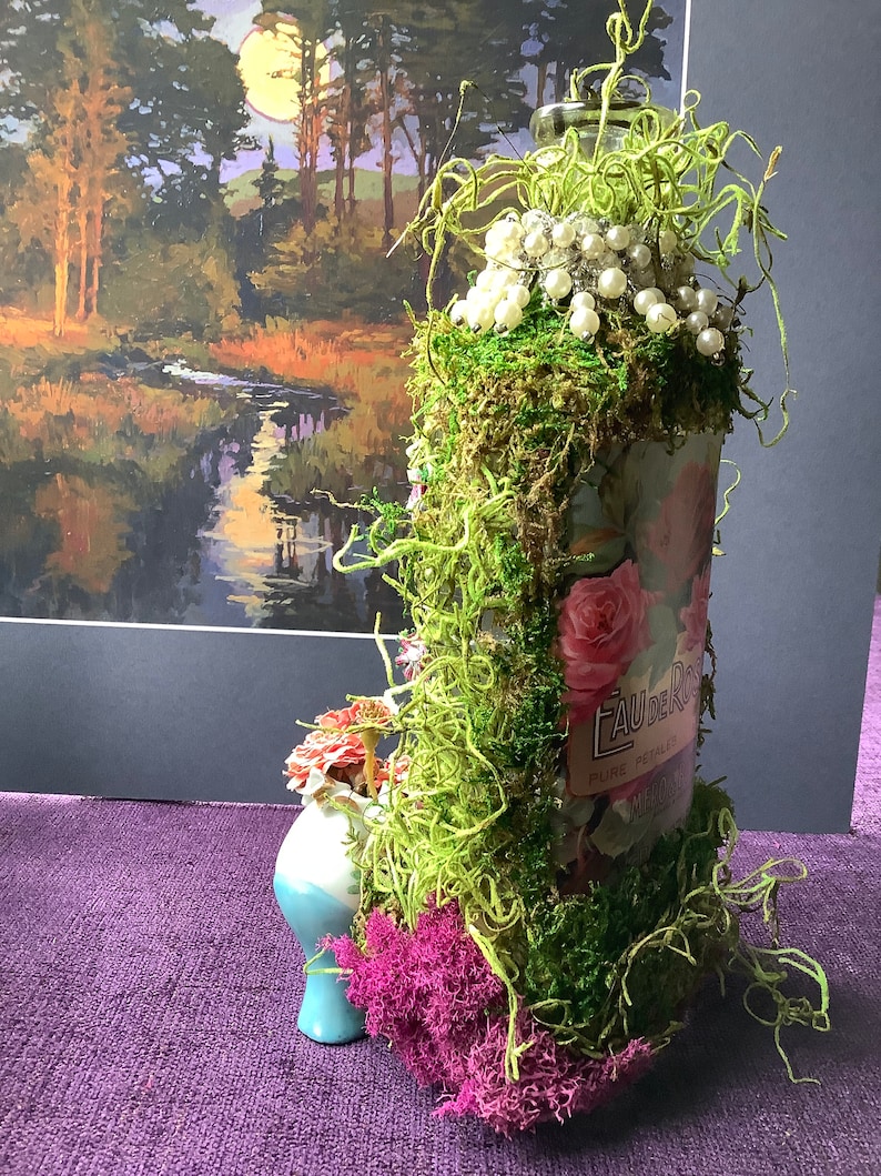 May include: A glass bottle decorated with moss, pearls, and a floral design. The bottle has a label that reads "Eau de Rose, Pure Petals".