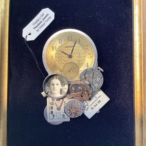May include: A framed mixed media artwork featuring a vintage clock face, watch parts, and a small photograph of a young girl. The artwork is titled "Daddy's Girl Timeless Jewels by Cheryl Young".