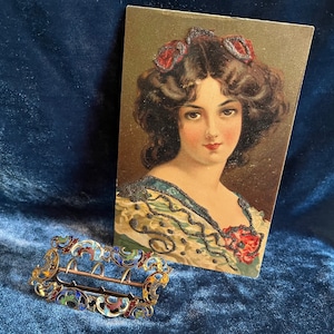 Op de afbeelding: Een vintage ansichtkaart met een vrouw met donker haar en een bloemenjurk. Ze draagt een juwelen hoofdband en heeft een serieuze uitdrukking. De ansichtkaart is in goede staat met wat slijtage aan de randen.