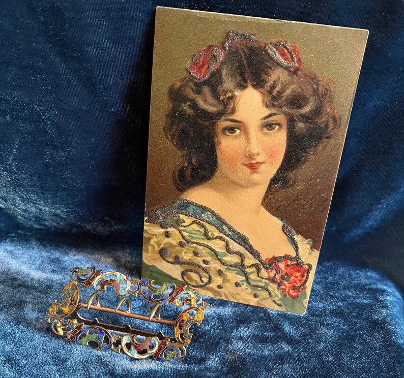 Op de afbeelding: Een vintage ansichtkaart met een vrouw met donker haar en een bloemenjurk. Ze draagt een juwelen hoofdband en heeft een serieuze uitdrukking. De ansichtkaart is in goede staat met wat slijtage aan de randen.