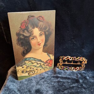 Op de afbeelding: Een vintage ansichtkaart met een portret van een vrouw met donker haar en een rode bloem in haar haar. De vrouw draagt een groene en zwarte jurk met een parelmoerontwerp. De ansichtkaart is in goede staat en heeft een lichte verkleuring aan de randen.