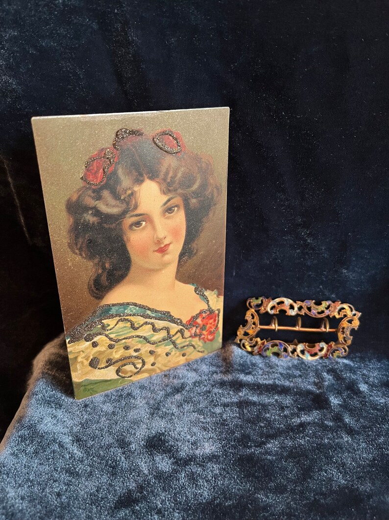 Op de afbeelding: Een vintage ansichtkaart met een portret van een vrouw met donker haar en een rode bloem in haar haar. De vrouw draagt een groene en zwarte jurk met een parelmoerontwerp. De ansichtkaart is in goede staat en heeft een lichte verkleuring aan de randen.