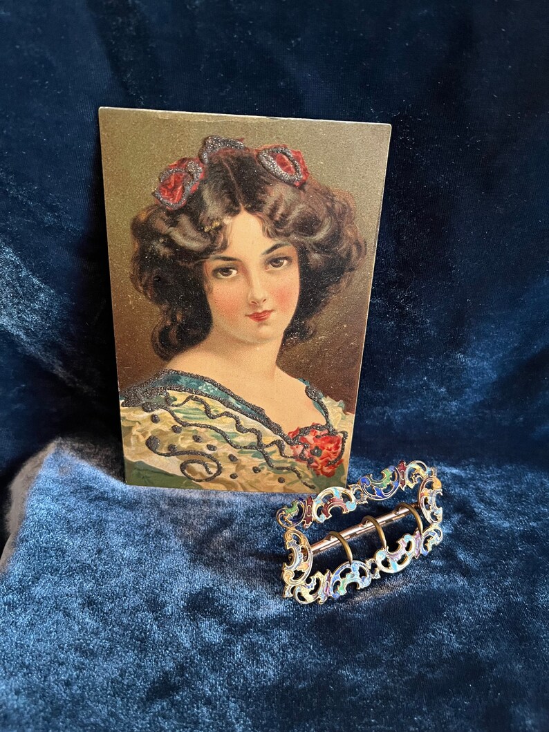 Op de afbeelding: Een vintage ansichtkaart met een vrouw met donker haar en een rode strik, naast een gouden gesp met blauwe, groene en rode emaille accenten.