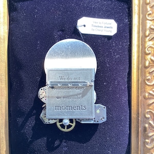 May include: A silver metal framed artwork with a quote that reads "We do not remember days, we remember moments". The artwork is decorated with gears and other metal pieces. The artwork is framed in a gold frame and has a tag that reads "Heir to Fortune Timeless Jewels by Cheryl Young".