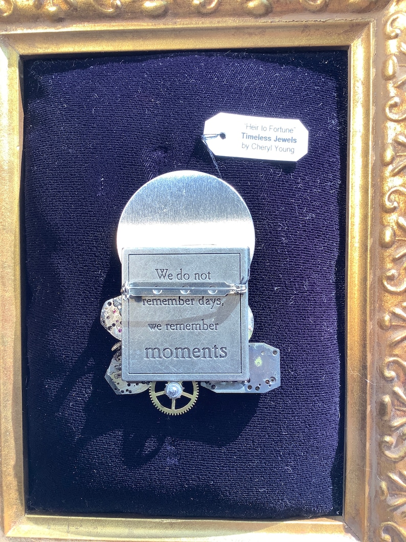 May include: A silver metal framed artwork with a quote that reads "We do not remember days, we remember moments". The artwork is decorated with gears and other metal pieces. The artwork is framed in a gold frame and has a tag that reads "Heir to Fortune Timeless Jewels by Cheryl Young".