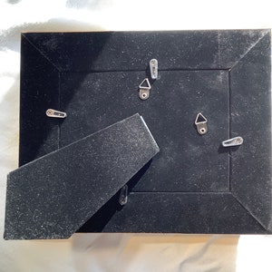 May include: The back of a black picture frame with a black velvet backing. The frame has four metal hanging hooks.