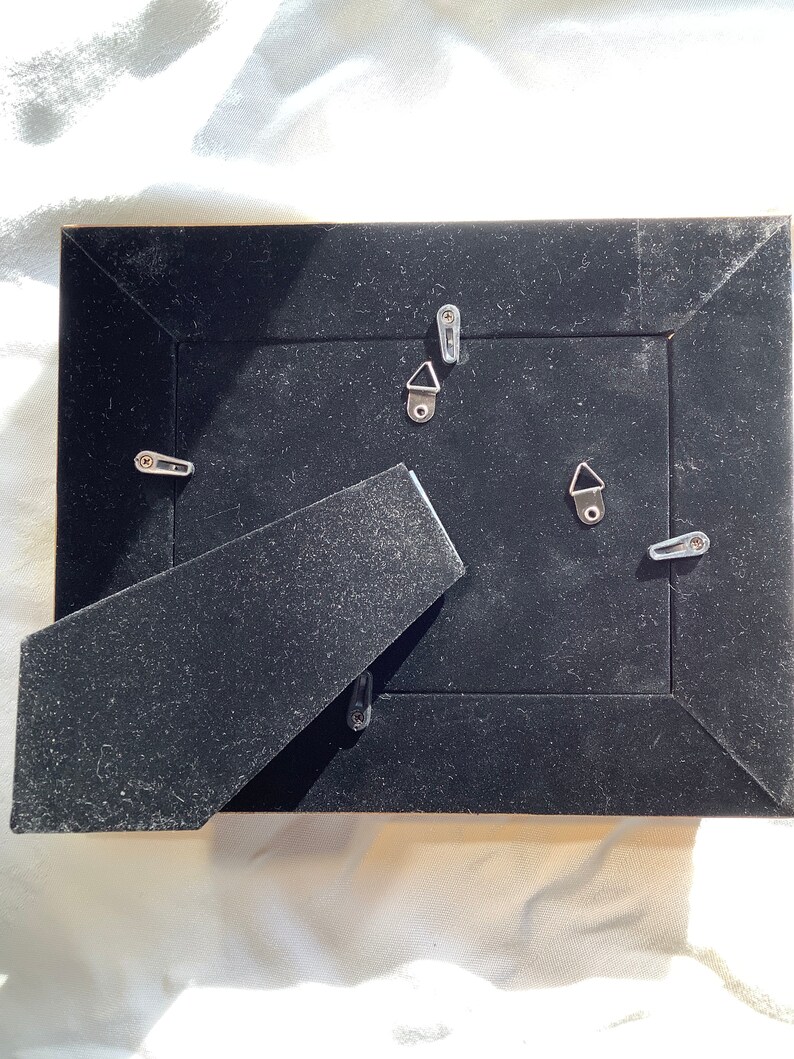 May include: The back of a black picture frame with a black velvet backing. The frame has four metal hanging hooks.