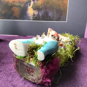 May include: A blue porcelain shoe figurine with floral details, nestled in a bed of green moss and pink flowers. The shoe is resting on a clear glass bottle with a red inscription. The figurine is made in Japan.