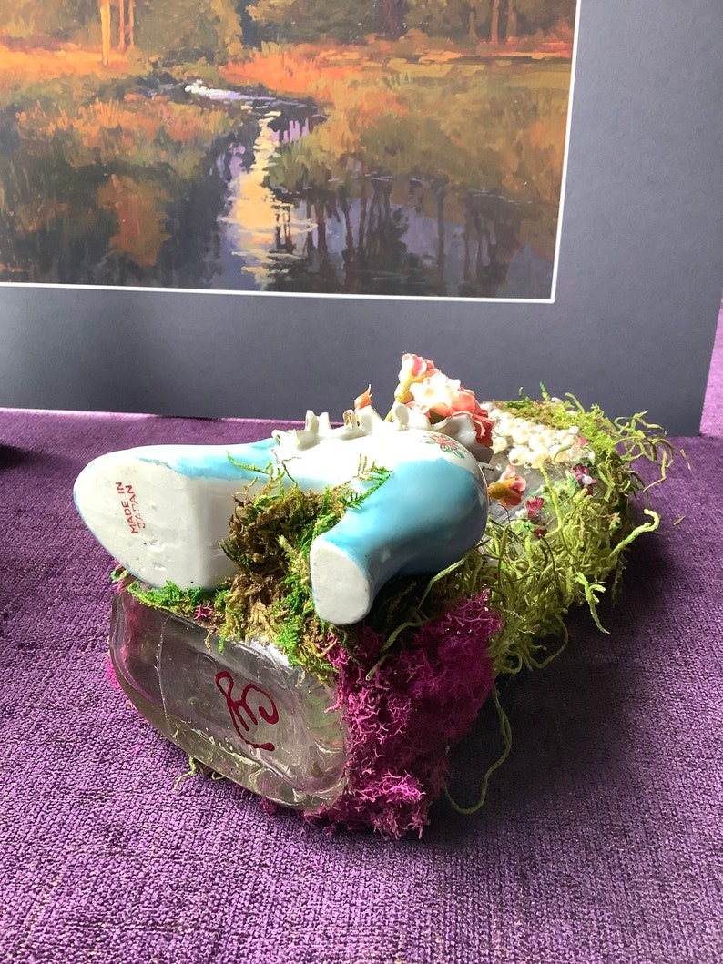 May include: A blue porcelain shoe figurine with floral details, nestled in a bed of green moss and pink flowers. The shoe is resting on a clear glass bottle with a red inscription. The figurine is made in Japan.