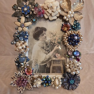 May include: A white rectangular picture frame adorned with vintage brooches in various shapes and colors, including blue, white, and gold. The frame surrounds a black and white photograph of a woman holding flowers.