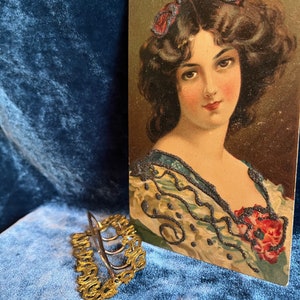 Op de afbeelding: Een gouden haarspeld met een decoratief ontwerp. De speld ligt op een blauw fluwelen oppervlak. Op de achtergrond is een vintage ansichtkaart te zien van een vrouw met donker haar en een blauw-witte jurk.